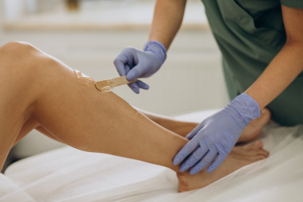 Esteticista com luvas azuis aplicando cera na perna de uma cliente para depilação, em um ambiente de spa ou clínica de estética.