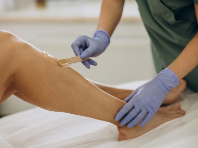 Esteticista com luvas azuis aplicando cera na perna de uma cliente para depilação, em um ambiente de spa ou clínica de estética.